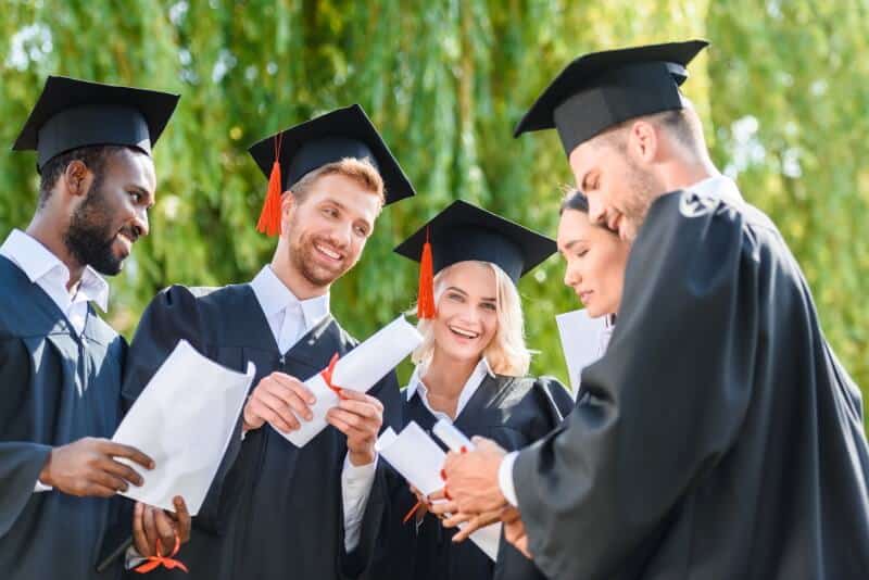 Five college graduates conversing wearing caps and gowns and holding diplomas