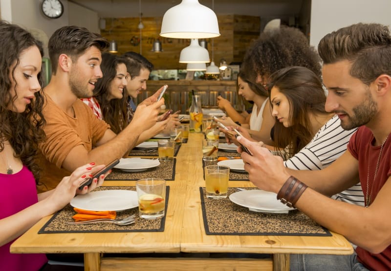 Group of young men and women at a table in a restaurant. All of them are looking at their mobile devices.